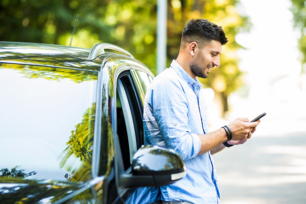 A driver, standing next to his car on a sunny day while he checks his phone.