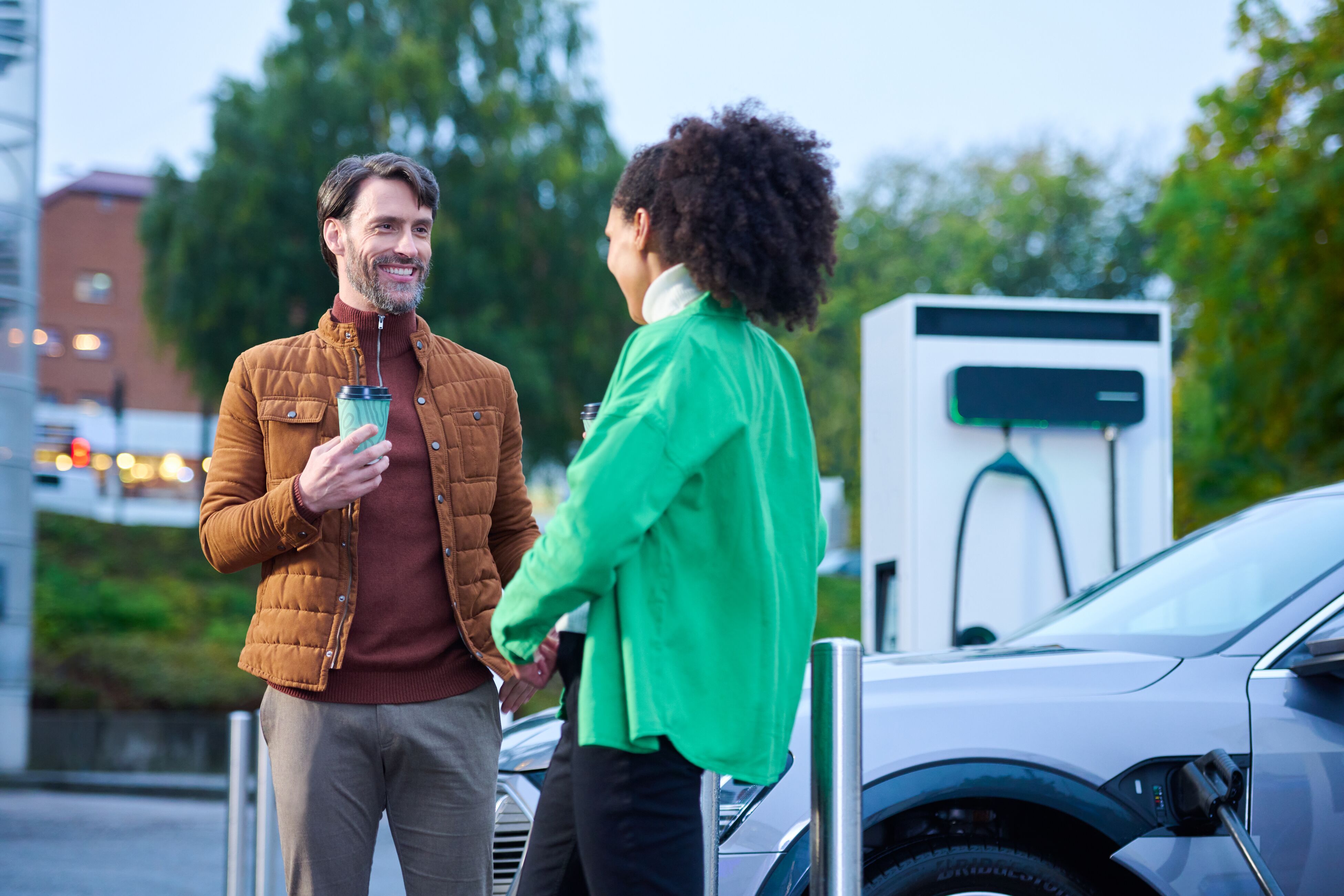 Man and woman talking next to electric car and EVBox Troniq Modular charging station, while enjoying a cup of coffee.