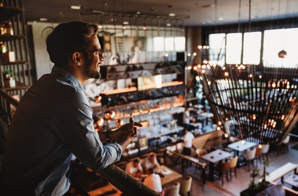 A restaurant owner smiling as he casually hangs across a first-floor balcony to look over his establishment.