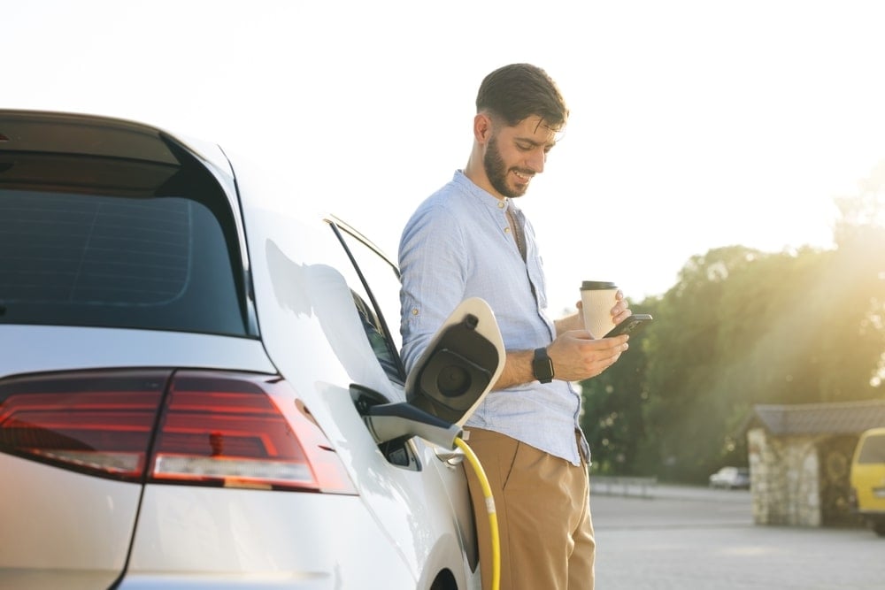 A man is waiting for his EV to be charged while holding a coffee cup.