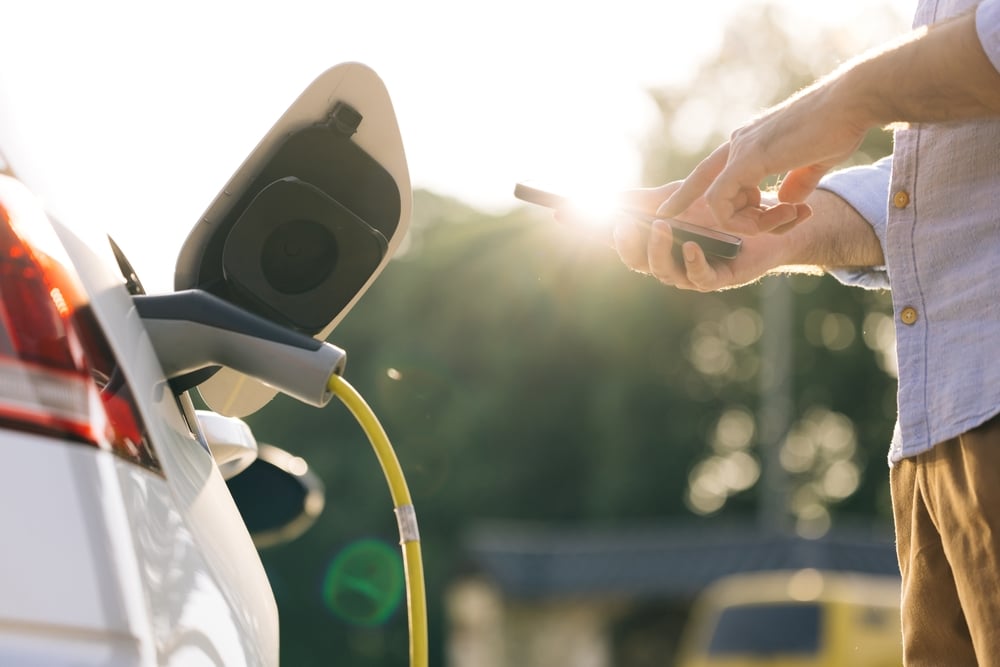 A man stands in front of his charging EV, holding his smart phone for real-time charging session insights.