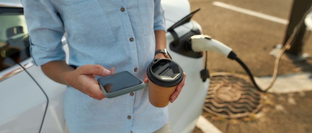 A woman charging her car while checking het phone and holding a take-away coffee cup.