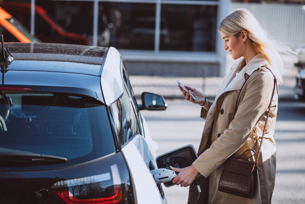 Woman charging electric car while looking at her smartphone.