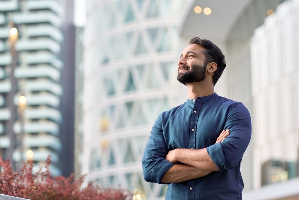 Man with crossed arms looks upwards with a soft smile. There are buildings and plants in the background.