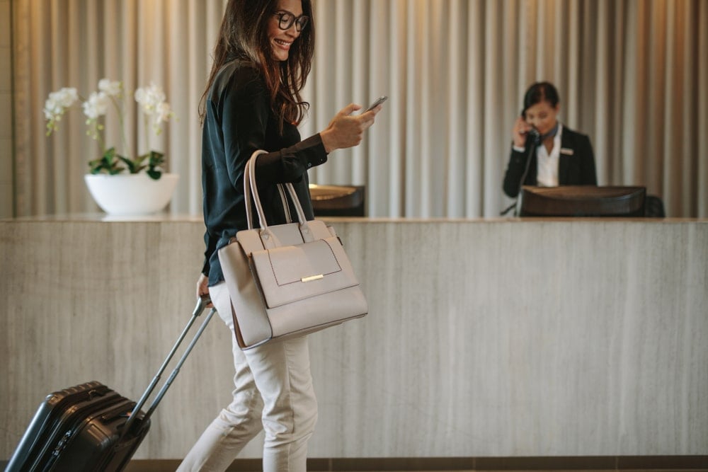 Woman carrying suitcase and a purse, smiling while looking at her cell phone, walks by the lobby of an upscale hotel.