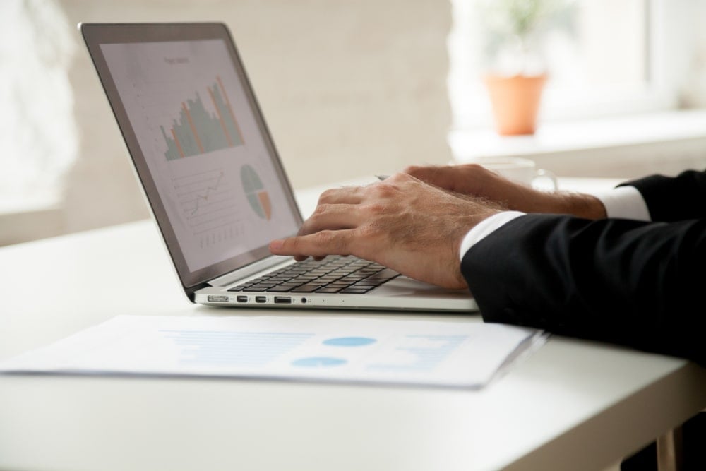 Man in suit typing on a laptop. There are graphs on the screen and on papers next to the device.