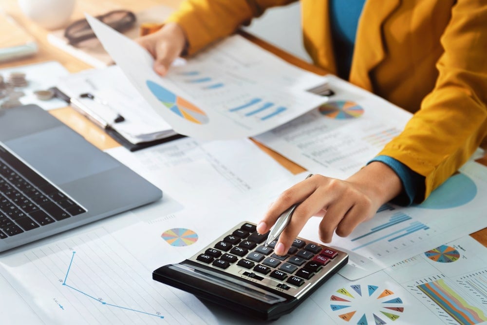 Hand using a calculator. There are papers with graphs on a table and a laptop.