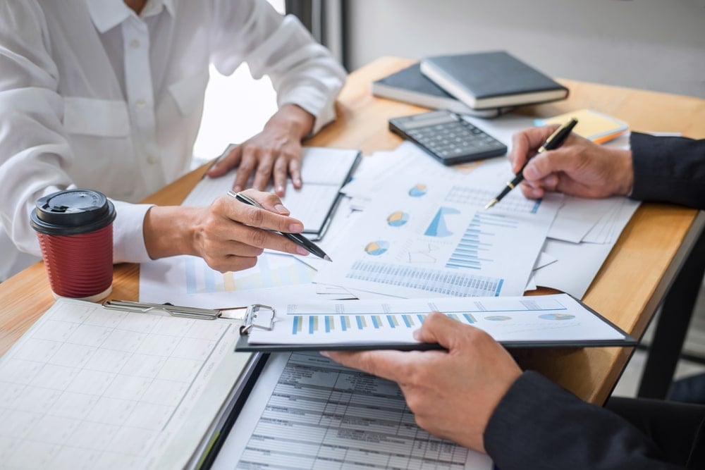 Two people pointing at graphs on papers on a table. There is also a calculator and a cup of coffee.